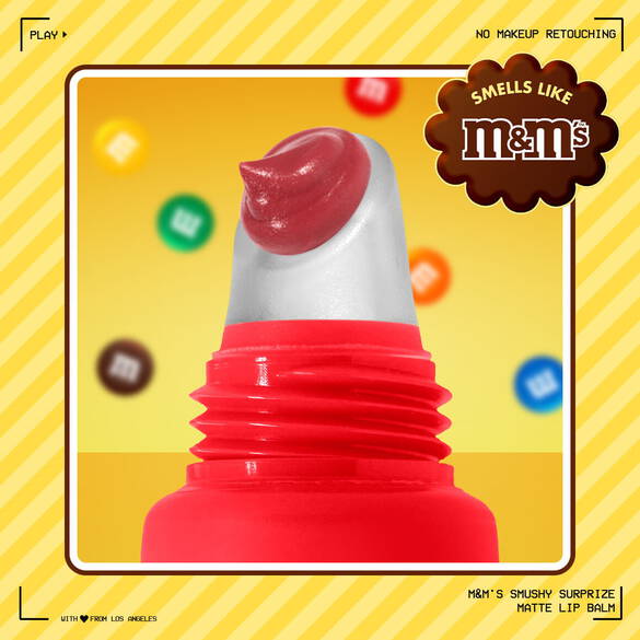 Primo piano del balsamo labbra opaco NYX Professional Makeup M&M’S Smushy Surprize con la punta dell'applicatore rossa, su uno sfondo giallo con colorati confetti M&M’S. Il testo mette in evidenza il profumo di M&M’S e l'assenza di fotoritocco sul makeup.