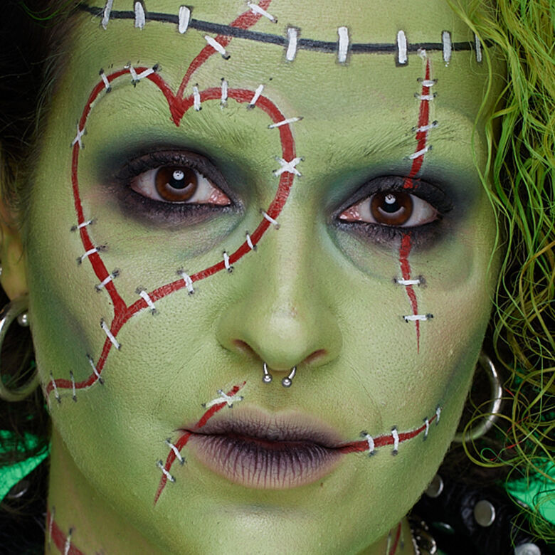 Look viso con cuciture verde di Halloween di NYX Professional Makeup con pittura pelle verde, linee di cucitura rosse, occhi scuri e labbra nere.