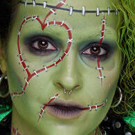 Look viso con cuciture verde di Halloween di NYX Professional Makeup con pittura pelle verde, linee di cucitura rosse, occhi scuri e labbra nere.