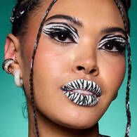 Look zebra di Halloween di NYX Professional Makeup con pittura viso bianca, strisce di zebra nere, eyeliner grafico e labbra a strisce.