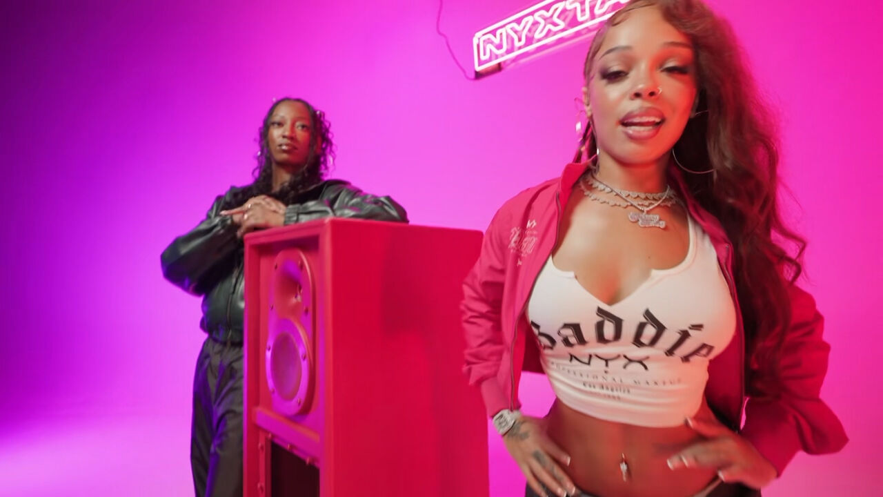 Scena del videoclip musicale di 'NYXTAPE' di NYX Professional Makeup con Wondagurl e Honey Bxby accanto a una cassa acustica rossa in uno studio illuminato di rosa, con un'insegna al neon 'NYXTAPE' in alto.