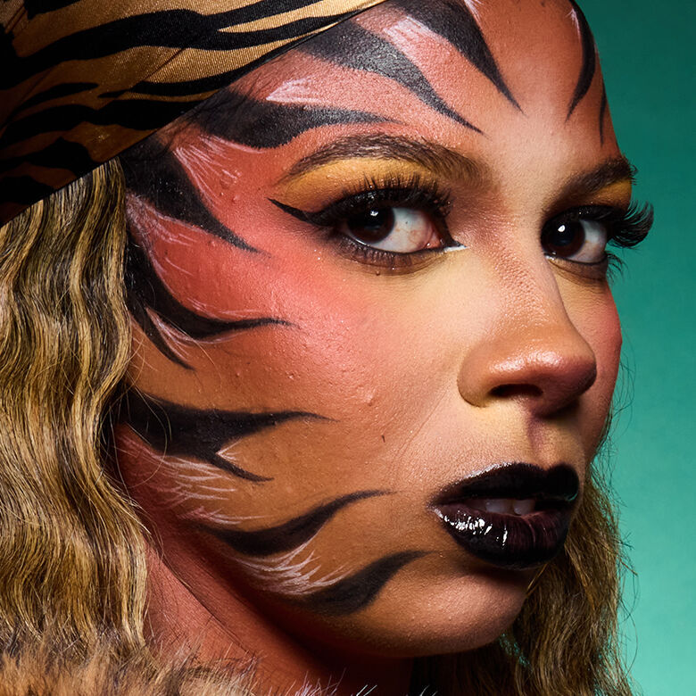 Look tigre di Halloween di NYX Professional Makeup con pittura viso arancione, strisce di tigre nere, trucco occhi oro e labbra nere lucide.
