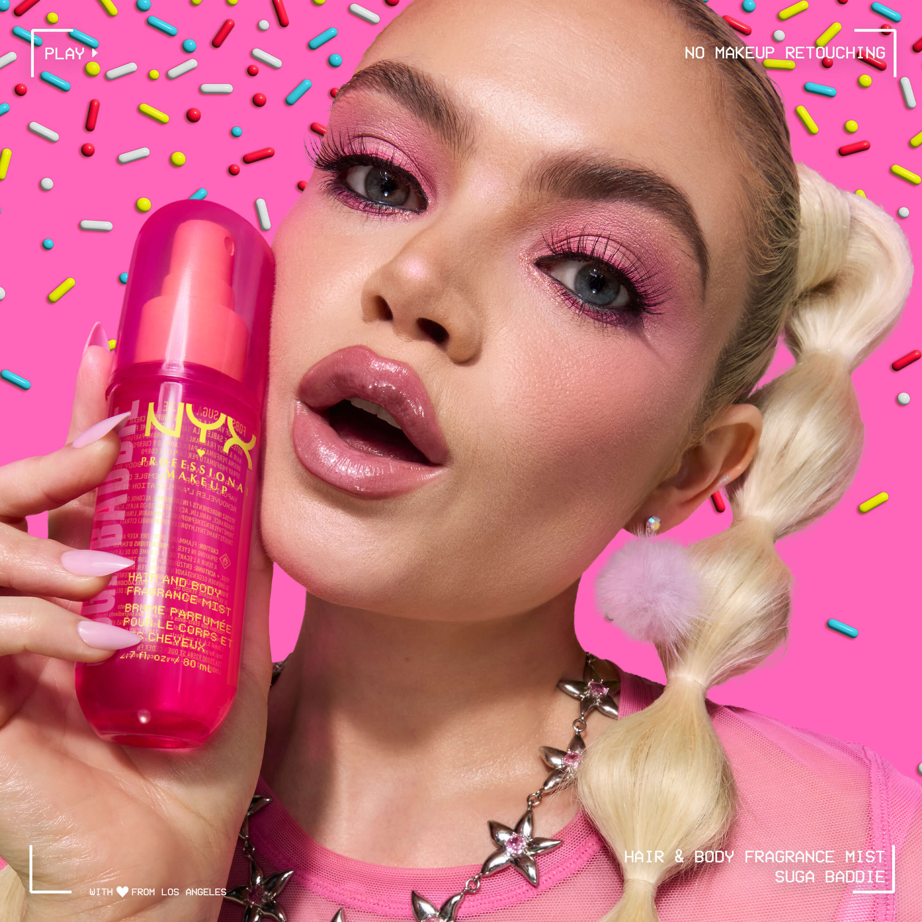 La modella tiene lo spray profumato capelli e corpo NYX Professional Makeup Suga Baddie in un flacone rosa acceso su uno sfondo rosa decorato con zuccherini colorati.