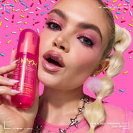 La modella tiene lo spray profumato capelli e corpo NYX Professional Makeup Suga Baddie in un flacone rosa acceso su uno sfondo rosa decorato con zuccherini colorati.