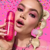 La modella tiene lo spray profumato capelli e corpo NYX Professional Makeup Suga Baddie in un flacone rosa acceso su uno sfondo rosa decorato con zuccherini colorati.