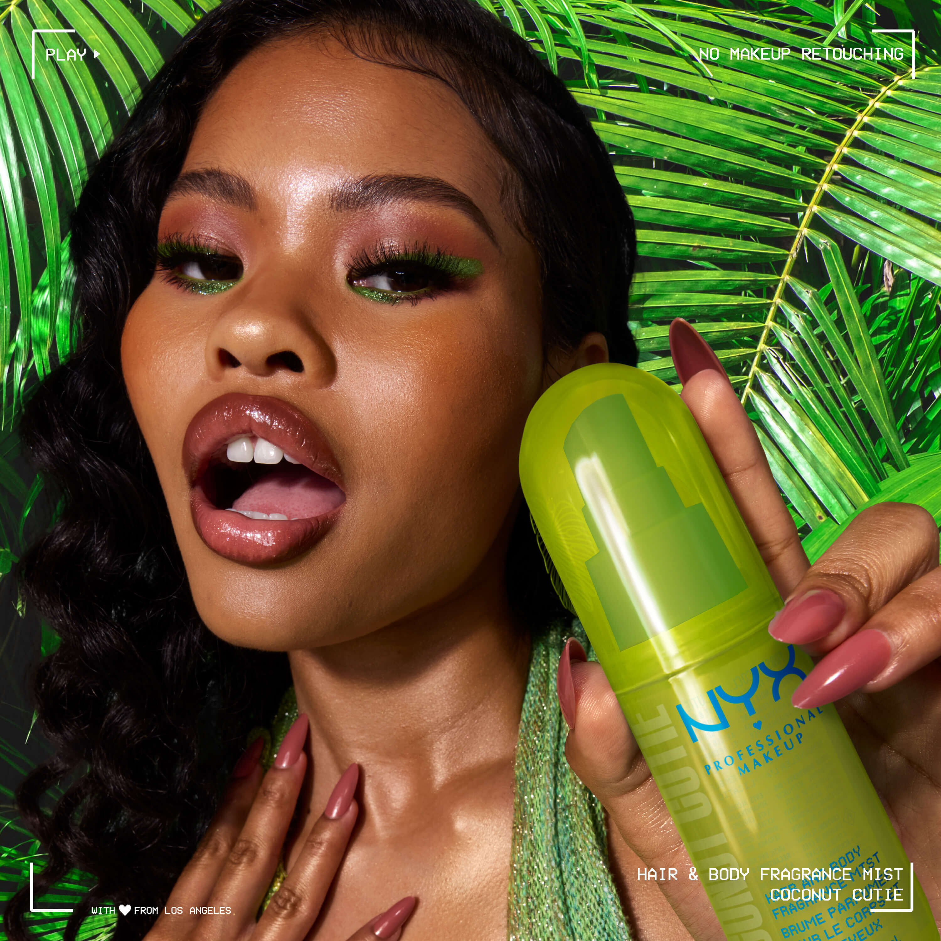 La modella tiene lo spray profumato capelli e corpo NYX Professional Makeup Coconut Cutie in un flacone verde neon su uno sfondo di foglie di palma tropicali