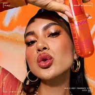 La modella tiene lo spray profumato capelli e corpo NYX Professional Makeup Juicy Boo in un flacone arancione acceso su sfondo arancione.
