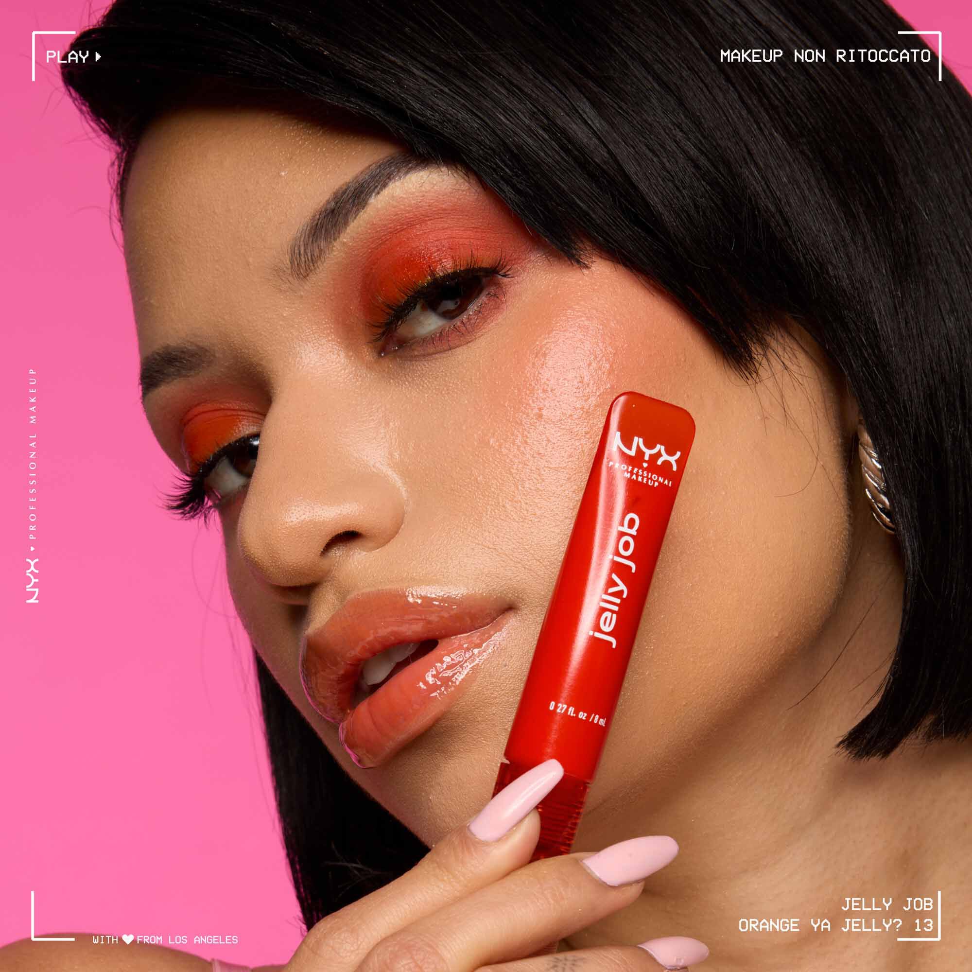 Close-up of a model with glossy lips, wearing red-toned eyeshadow and holding a red NYX Jelly Job lip gloss. Background is pink. Text reads: "Jelly Job - Orange Ya Jelly? 13".-3